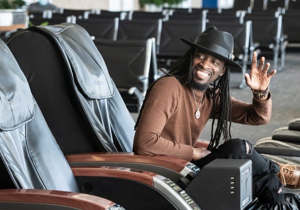 Man wearing a fedora smiling and waving as he sits in a massage chair.
